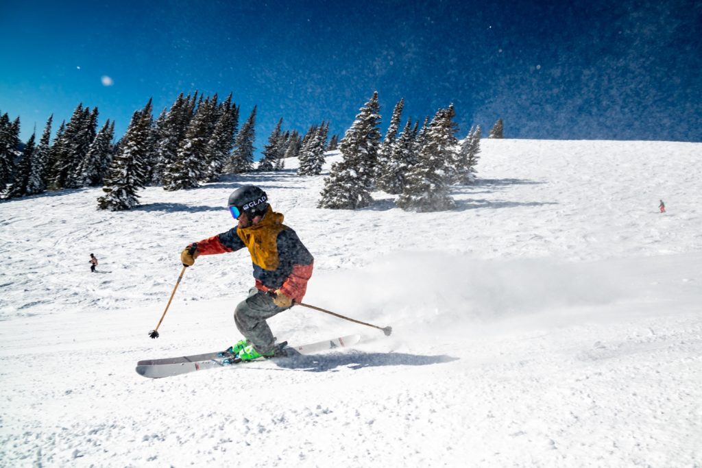 Ein Skifahrer auf einer weißen Piste vor einem Walt und wunderschönen blauen Himmel.