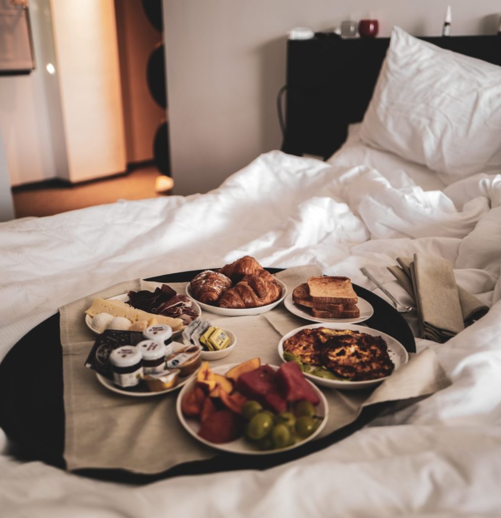 Ein Tablett steht auf einem Bett mit weißer Bettwäsche. Am Tablett befinden sich verschiedene leckere Frühstücksspeisen.